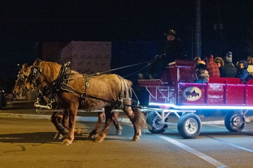 A horse-drawn sleigh with a bright red carriage glides through a nighttime street, carrying several passengers bundled in winter clothing. The sleigh features a bat logo and is illuminated with lights, adding a festive touch to the scene.