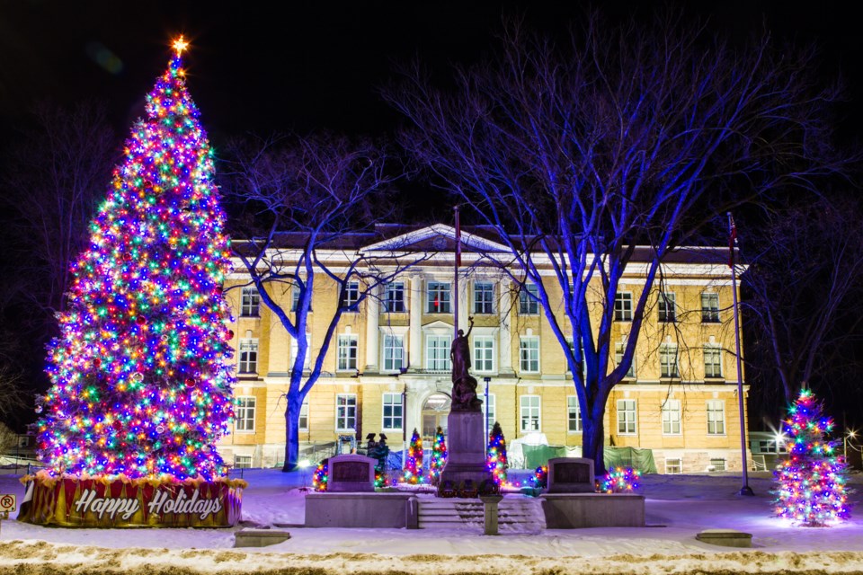 A brightly lit Christmas tree adorned with colorful lights stands in front of a historical building, with snow on the ground. The scene is festive and inviting, showcasing holiday decorations and a warm ambiance at night.