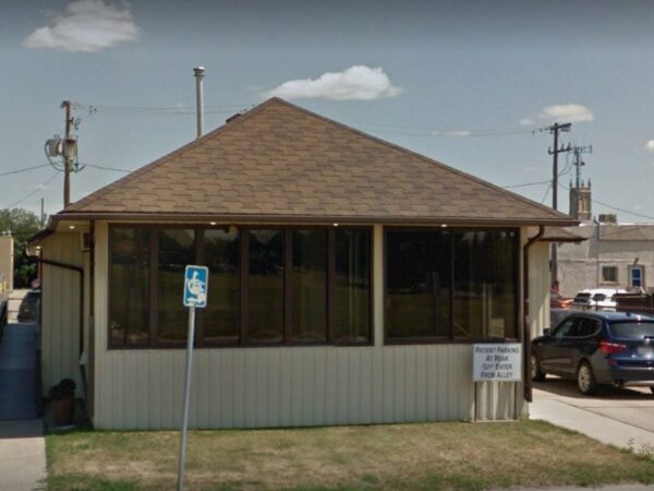 A small, one-story building with a sloped roof and large front windows, surrounded by grassy areas. A ramp leads to the entrance, and there are parking spaces visible in the foreground. Utility poles and a distant church tower can be seen in the background.