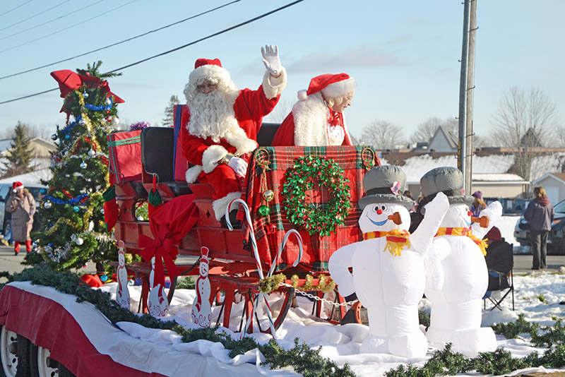 Two Santa Claus figures wave from a festive sleigh adorned with greenery and decorations, accompanied by cheerful snowman figures in a snowy parade setting.