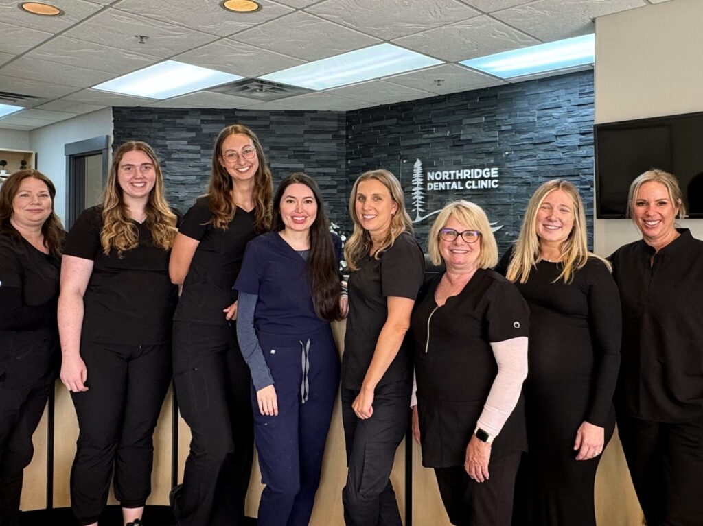 A group of eight dental professionals stands together in a clinic, wearing black attire. They display friendly smiles, with a welcoming ambiance highlighted by the clinic's modern decor. The 123Dentist Smiley is joyfully featured, adding a cheerful touch to the scene.