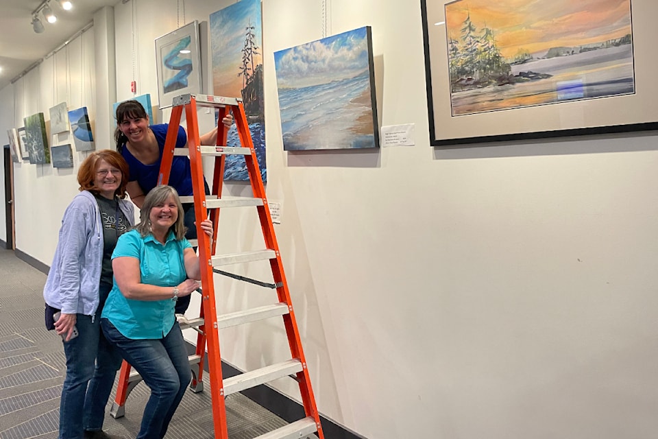 Three women smile in an art gallery as they arrange paintings on the wall. One woman stands on a ladder, while the others pose beside it. The walls are decorated with various landscapes.