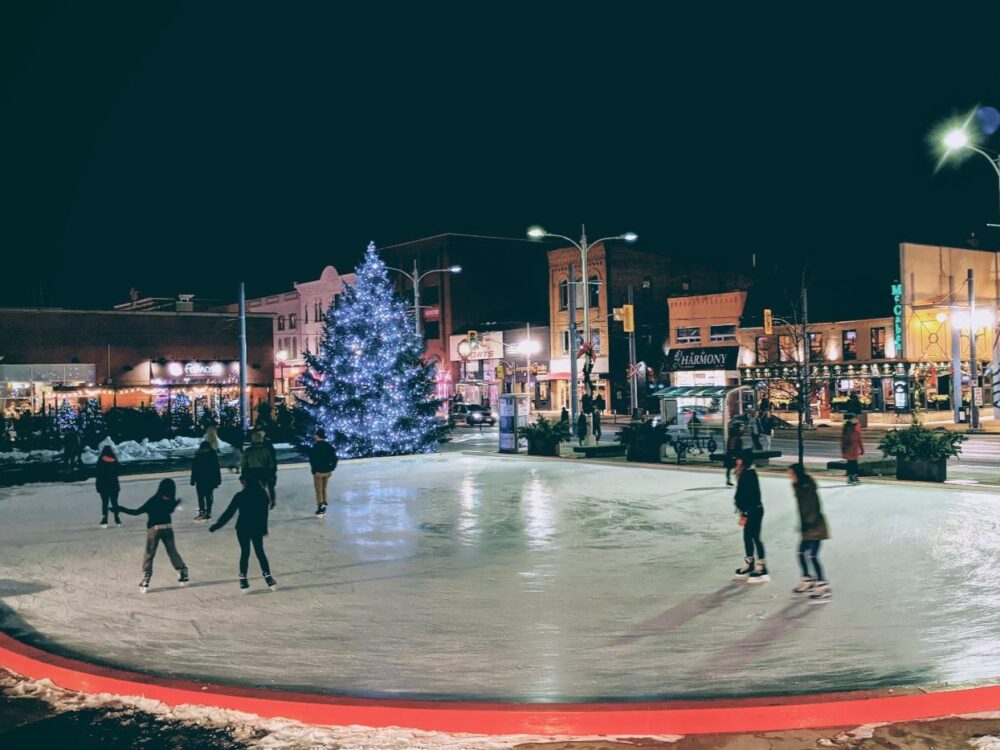 A festive outdoor ice skating rink at night, surrounded by illuminated buildings and a large, decorated Christmas tree. Skaters enjoy the icy surface, creating a joyful winter atmosphere.