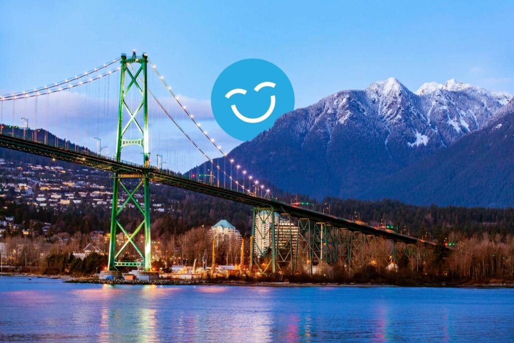 A colorful suspension bridge spans a river, with snow-capped mountains in the background. A cheerful, smiling icon overlays the scene, adding a vibrant touch to the landscape at dusk.
