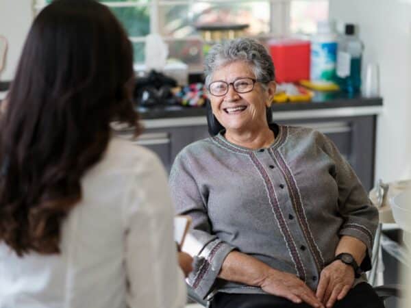 Une femme souriante d'un certain âge est assise dans un cabinet dentaire et s'entretient avec un professionnel de la santé. Le cadre est lumineux et accueillant, reflétant une atmosphère amicale. L'émoticône 123Dentiste ajoute une touche de gaieté à la scène, favorisant une expérience dentaire positive.