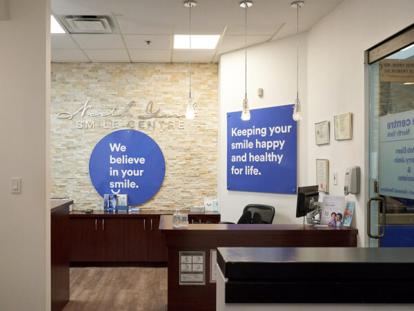 A dental office reception area featuring a welcoming desk, soft lighting, and motivational wall signs about smiles. The blue circle sign states, "We believe in your smile," while another emphasizes keeping smiles happy and healthy for life. The setting conveys a friendly and professional atmosphere.