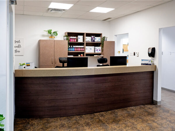 A modern dental office reception area featuring a wooden front desk, plants, and organized shelves with dental products. The space is bright and welcoming, designed to create a comfortable atmosphere for patients.