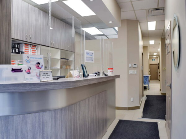 A modern reception area featuring a wooden front desk with protective screens, organized office supplies, and informational signage. Soft lighting illuminates the space, leading down a clean hallway with visible doors, creating a welcoming and orderly environment.