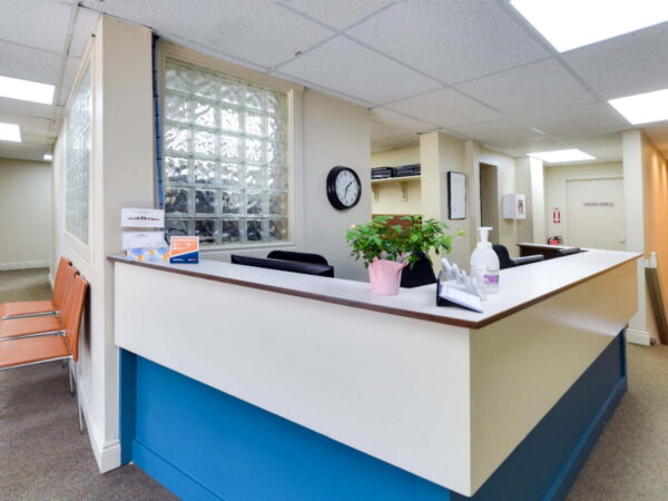 A reception area featuring a desk with a clock, potted plants, and seating. The space is well-lit and includes a window and beige walls, creating a welcoming atmosphere for visitors.