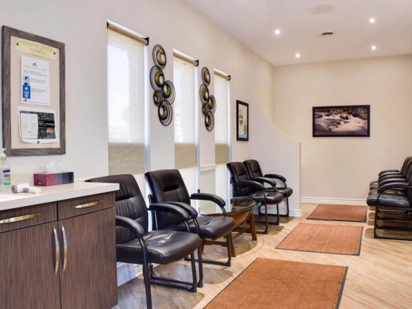 A waiting area featuring several black chairs, a small table, and two rugs. Light-colored walls are decorated with framed artwork and circular wall hangings. Natural light enters through window shades.