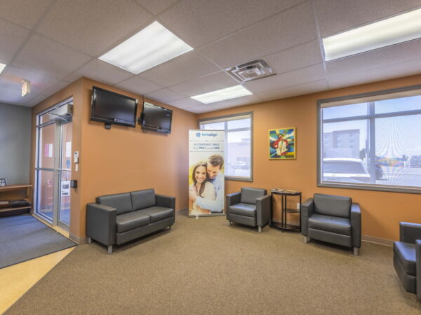 A modern waiting area featuring gray seating, a large window providing natural light, and a decorative wall art piece. Two televisions are mounted on the wall, and a banner stands nearby, enhancing the inviting atmosphere. The space is clean and well-organized.