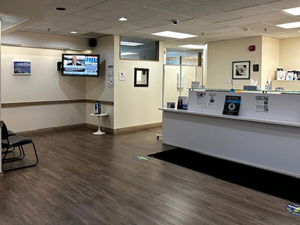 A modern dental office reception area featuring a clean, well-lit space with a waiting area, a reception desk, and informational displays. A television is mounted on the wall, providing entertainment for patients, while the 123Dentist Smiley adds a friendly touch to the environment.
