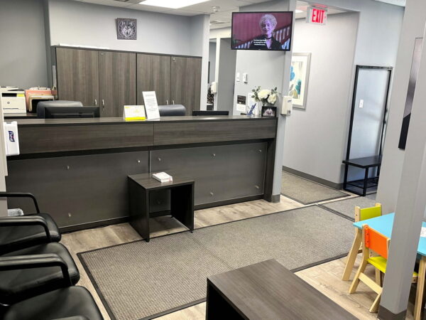 A modern office reception area featuring a front desk, multiple chairs, a small children's play table, and a television on the wall. The space is well-lit with gray and wooden accents, creating a welcoming environment for visitors.