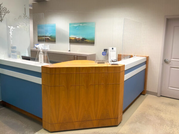 A modern dental reception area featuring a wooden and blue reception desk, equipped with a clear protective barrier. The wall displays serene landscapes in artwork and a door leads to another room, creating a welcoming atmosphere for patients.