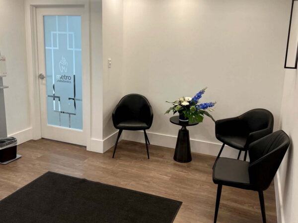 A modern waiting room featuring two black chairs and a small round table with a floral arrangement. A door with glass panels leads outside, and a minimalist design creates a welcoming atmosphere. A black mat is on the floor, enhancing the overall aesthetic.