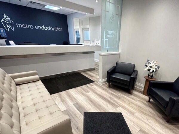 A modern dental office waiting area featuring a white reception desk, a light-colored tufted sofa, and two black chairs. The walls are painted light gray, and there is a decorative plant on a small table. The sign "metro endodontics" is prominently displayed.