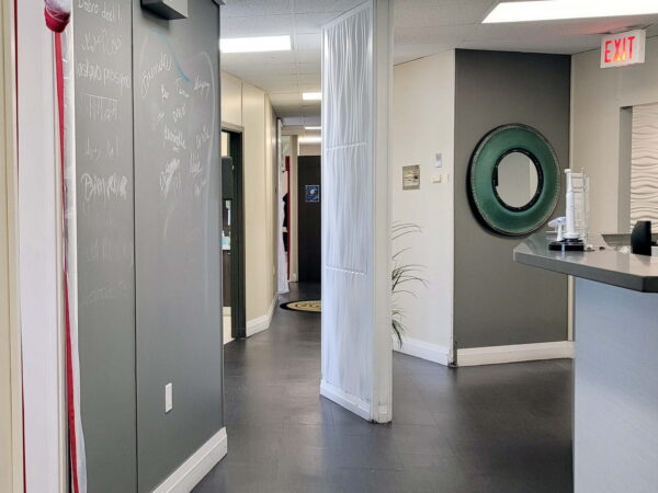 A well-lit hallway in a modern office setting, featuring gray walls, a reception desk, and a round mirror. The space is organized with a comfortable ambiance, leading to various rooms and areas.
