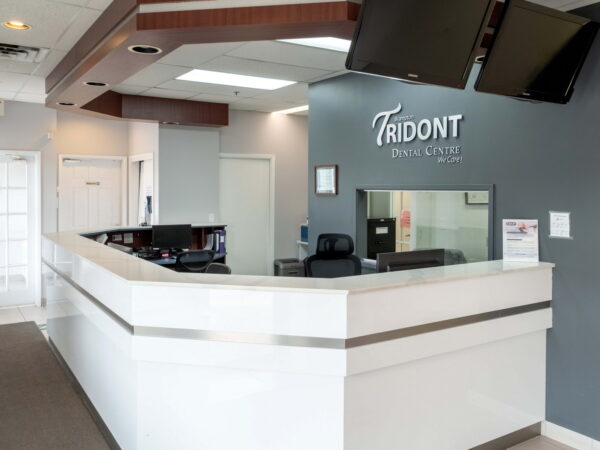 A modern dental clinic reception area featuring a sleek white countertop, gray walls, and a welcoming atmosphere. The Tridont Dental logo is prominently displayed, with seating and informational materials visible in the background.