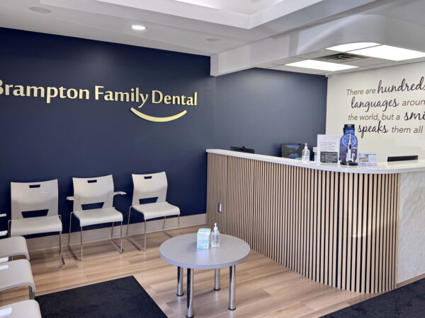 A modern dental office reception area featuring a dark blue wall with the text "Brampton Family Dental," light-colored wooden reception desk, seating for patients, and minimal decorative elements promoting a welcoming atmosphere.