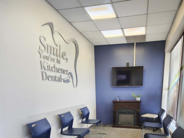 A waiting room for a children's dental office featuring blue walls with a large decorative text saying, "Smile, You're at Children's Dental." There are several blue chairs arranged, a television on the wall, and a decorative element like a small plant.