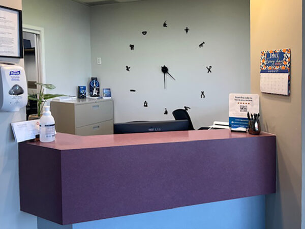 A dental reception area featuring a purple countertop, a wall clock with a minimalist design, and an organized workspace. There is a calendar and informational materials visible, along with hand sanitizer on the counter. A friendly blue 123Dentist Smiley is present, adding a welcoming touch.