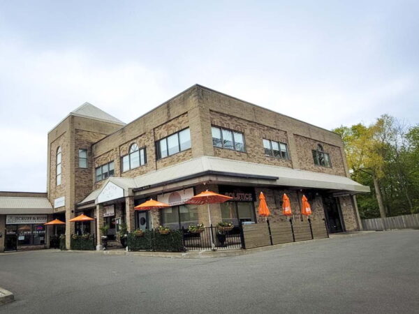 A modern brick building with large windows, featuring an outdoor seating area with orange umbrellas. The structure is situated in a landscaped area, indicating it could be a restaurant or café, set against a cloudy sky.