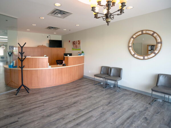 A modern reception area featuring a wooden front desk, two chairs, a coat rack, and a round mirror. The space is well-lit with neutral colors and wooden flooring, creating a welcoming atmosphere.
