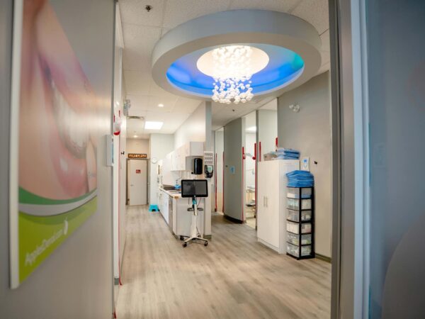 A bright and modern dental office hallway featuring a chandelier, light blue ceiling, and sleek furniture. The space is clean and inviting, with dental equipment and supplies visible in the background.