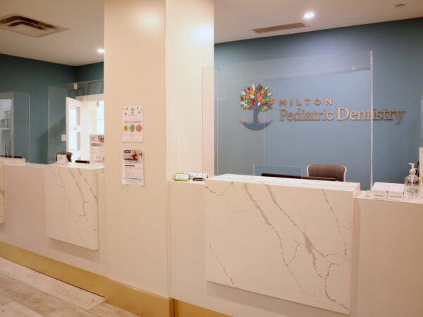A modern dental reception area featuring a clean, white marble front desk with a logo of a tree. The walls are painted blue, and safety barriers are visible. Informational signs are displayed on the side, creating a welcoming and professional atmosphere for patients.