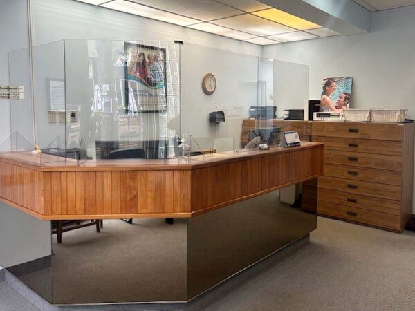 A reception area featuring a wooden front desk with a glass partition, a filing cabinet, wall art, and overhead lighting. The space is designed for customer interaction and is tidy with minimal decoration.