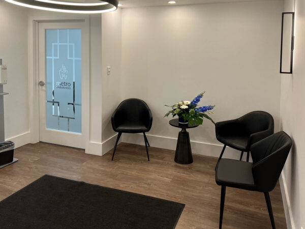 A modern dental office waiting area featuring two black chairs, a round table with flowers, and a stylish entrance door. Soft lighting enhances the inviting atmosphere, making it a comfortable space for patients. The friendly 123Dentist Smiley adds a touch of warmth to the environment.