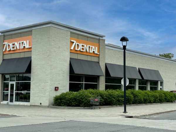 A modern dental office building with a large sign reading "7 DENTAL" is shown. The structure features large windows and awnings, surrounded by greenery, under a clear blue sky.