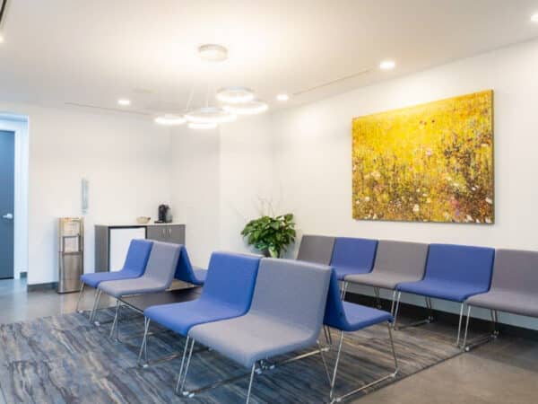 A modern dental waiting room featuring blue chairs arranged around a table. The walls are brightly lit, adorned with a large yellow artwork, and a potted plant adds a touch of greenery. The space is clean and inviting.