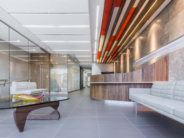 A modern office reception area featuring sleek furniture, a glass partition, and a wooden reception desk. The space is well-lit, with a stylish ceiling design and comfortable seating.