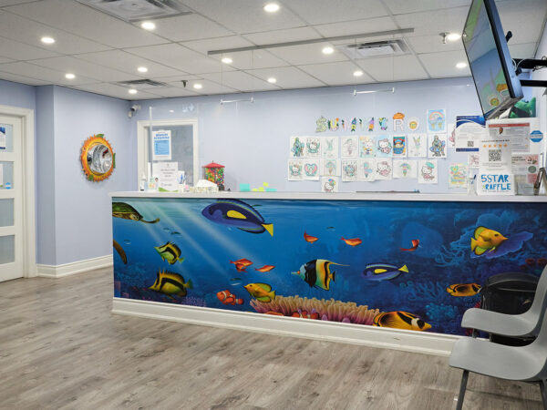 A bright dental office reception features a vibrant underwater mural with colorful fish. The space includes a welcoming desk, a television, and a chair for visitors. The light blue walls and cheerful decor create a friendly environment, perfect for easing patient anxiety. The 123Dentist Smiley adds a playful touch.