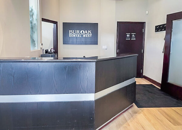 A modern dental office reception area featuring a sleek counter with a sign reading "Bur Oak Dental West." The space is warmly lit with wooden flooring, creating an inviting atmosphere for patients.