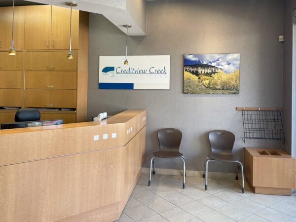 A dental office reception area featuring a wooden front desk, modern seating, and a landscape painting on the wall. Brightly lit and inviting, it provides a warm atmosphere for patients. The friendly 123Dentist Smiley adds a cheerful touch to the environment.
