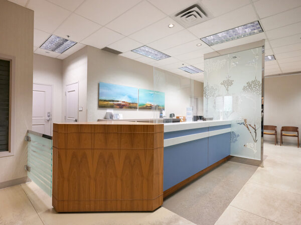 A modern dental office reception area featuring a wooden front desk with blue accents, glass partitions, and artistic wall decor. The space is well-lit, creating a welcoming atmosphere. The friendly 123Dentist Smiley adds a cheerful touch to the environment.