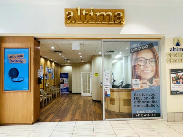 The image shows the entrance of an Altima dental clinic located in a shopping mall. The doorway features a warm wooden design and a promotional banner advertising free consultations, with seating visible inside the clinic.