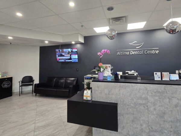 A modern dental clinic reception area featuring a dark wall with the "Airline Dental Centre" sign, a sleek front desk, a comfortable black sofa, and a television displaying information. Decor includes a floral arrangement and promotional materials on the counter.