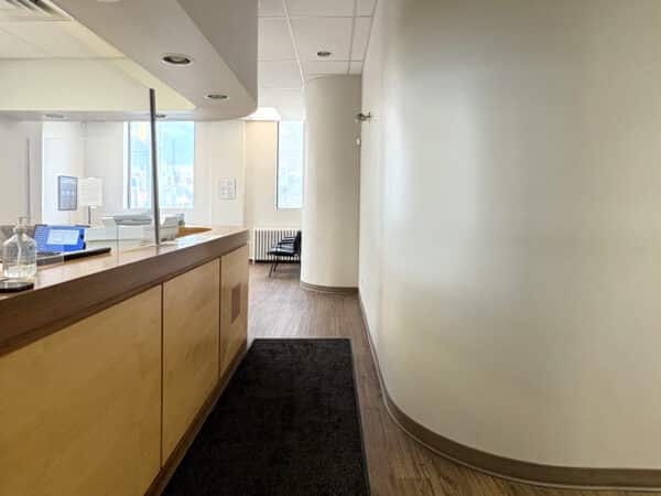 An inviting dental office corridor featuring a wooden reception desk, a warm ambiance, and natural light streaming through a large window. The space is neatly organized, leading to a comfortable waiting area. The friendly 123Dentist Smiley adds a welcoming touch.