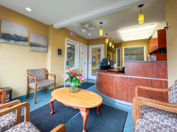 A bright and welcoming dental clinic waiting area featuring two patterned chairs, a wooden table with a flower vase, and a reception desk. Light-colored walls and artwork create a soothing atmosphere for patients.