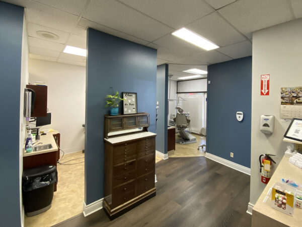 A bright dental office hallway features blue walls and wooden flooring. To the left, there is a reception area with furniture and plants. The space is inviting and well-organized, leading to dental rooms, creating a welcoming atmosphere for patients. 123Dentist Smiley adds a friendly touch to the environment.
