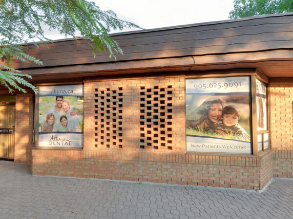 A dental office exterior featuring large windows with promotional posters displaying happy families and smiling faces. The building has a brick facade and is surrounded by greenery, creating a welcoming atmosphere.