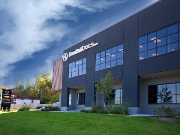 A modern dental clinic building named "DentalDocs," featuring large windows and a well-maintained grassy area. The sky is partly cloudy, adding to the inviting atmosphere of the scene.