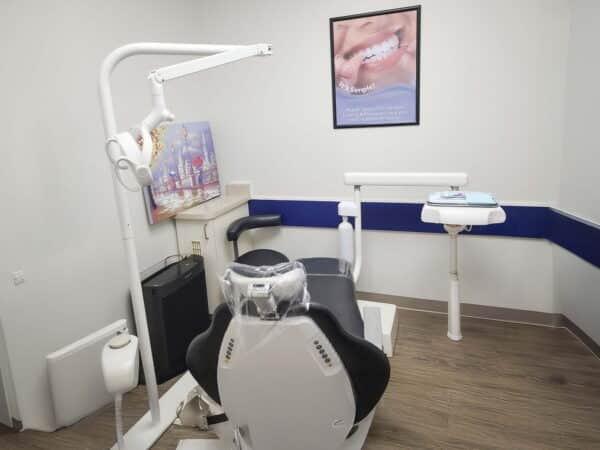 A dental clinic interior featuring a modern dental chair, overhead light, and adjacent treatment equipment. A framed picture hangs on the wall, adding to the clinical atmosphere. The floor is wood-colored, and the walls are accented with blue.