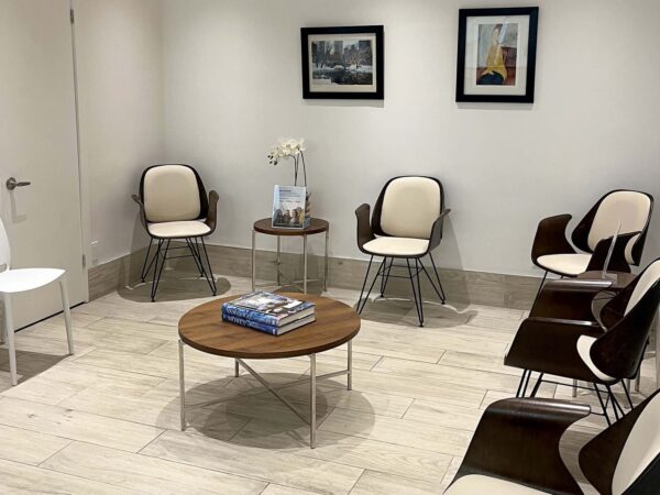 A modern waiting room featuring six chairs positioned around a central coffee table with a few magazines. The walls are adorned with two framed pictures, and a small decorative plant sits on the table, creating a clean and inviting atmosphere.