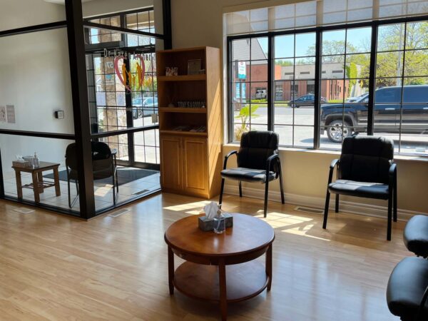 A bright waiting room featuring wooden flooring, a round coffee table, and several black chairs. Natural light streams through large windows, showcasing an outside view. A small bookshelf is positioned against one wall, adding a welcoming touch to the space.
