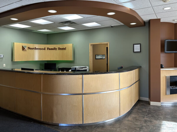 A welcoming dental office reception area featuring a curved wooden front desk, soft green walls, and a cozy atmosphere. The space includes seating and a wall-mounted television, designed to create a friendly and inviting environment for patients.