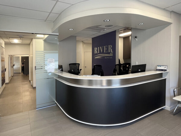 A modern dental office reception area featuring a sleek curved counter, a dark wall with the name "RIVER DENTAL," and comfortable seating nearby. The space is well-lit and organized, creating an inviting atmosphere for patients.